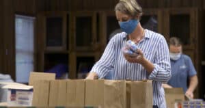 A woman stands over brown bags, filling them with food. She has her blue mask on to protect others around her from the spread of Covid-19.